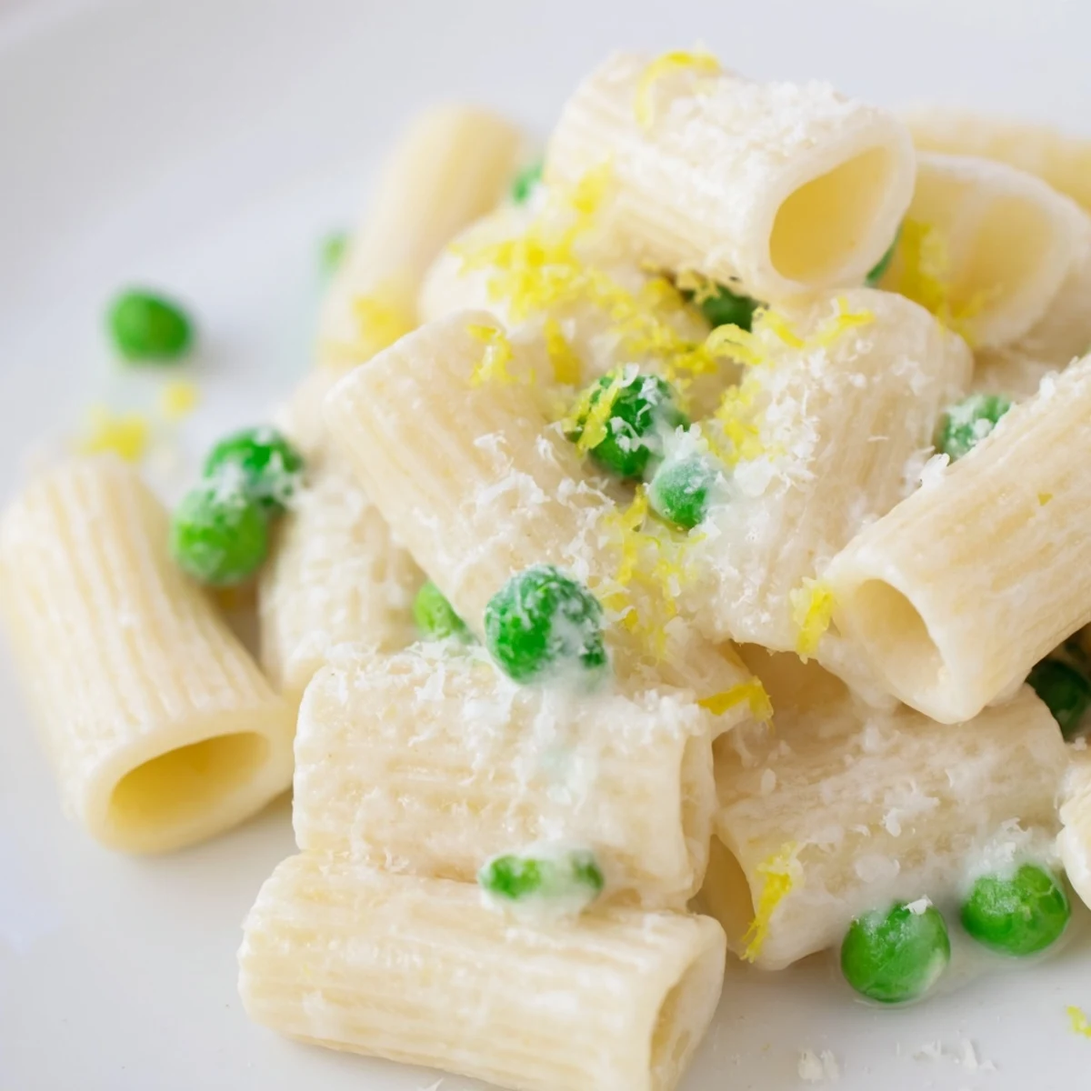 A close-up of Pea & Lemon Ricotta Pasta in a white bowl, featuring creamy ricotta sauce clinging to rigatoni and bright green peas, with fresh basil on top.